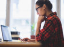 Mulher de camisa xadrez vermelha e óculos preto, com a mão no queixo, estudando na frente de laptop, estudando pra saber qual o perfil de investidor