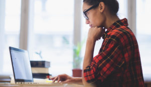 Mulher de camisa xadrez vermelha e óculos preto, com a mão no queixo, estudando na frente de laptop, estudando pra saber qual o perfil de investidor