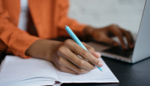 Mãos de mulher digitando em laptop e fazendo anotações sobre sua saúde financeira em um caderno