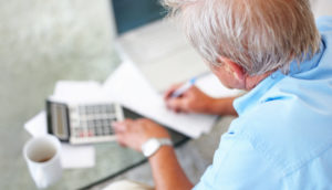 Pessoa sentada de costas fazendo contas com calculadora e a cartilha de educação financeira para idosos ao lado