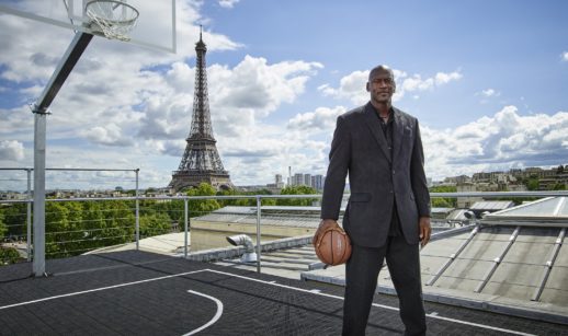 Michael Jordan de pé com terno, seu tênis, em quadra de basquete com fundo para a Torre Eiffel