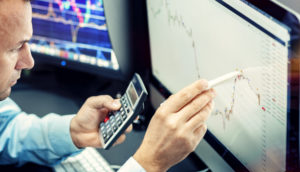 Especialista em frente ao computador com celular e caneta em mãos analisando ações para se investir em março