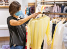 Mulher olhando blusa em loja de roupa com máscara. Loja vende fiado