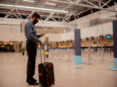 Homem com celular na mão, máscara e mala em aeroporto pronto para embarcar em avião