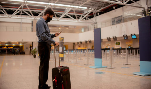 Homem com celular na mão, máscara e mala em aeroporto pronto para embarcar em avião