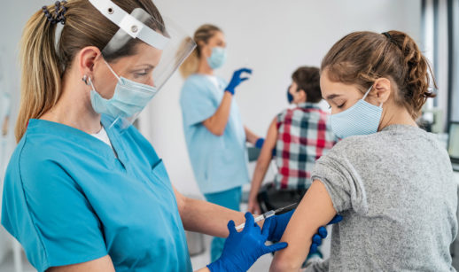 Enfermeira aplicando vacina da Pfizer em menina adolescentes, ambas de máscara
