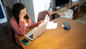 Mulher pensativa sentada em mesa com laptop, celular, papéis e caneta calculando IR 2021