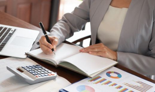 Pessoa com caneta e livro em branco, rodeada de papéis e calculadora, analisando balanços de empresas