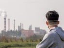 Homem de costas e máscara em primeiro plano olhando paisagem de chaminés de fábricas despejando gases do efeito estufa na atmosfera