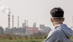 Homem de costas e máscara em primeiro plano olhando paisagem de chaminés de fábricas despejando gases do efeito estufa na atmosfera