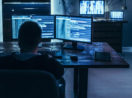 Imagem de hacker de costas, sentado em frente a várias telas e computador, planejando ataques contra os EUA