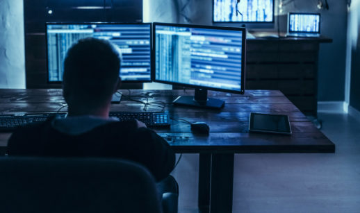 Imagem de hacker de costas, sentado em frente a várias telas e computador, planejando ataques contra os EUA