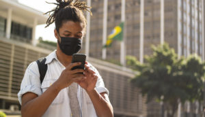 Homem usando celular de máscara na Avenida Paulista, em São Paulo, com a bandeira do Brasil de fundo