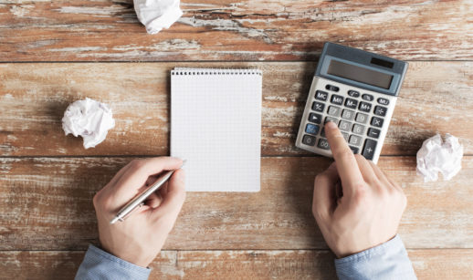 Mão de pessoa com papel, caneta e calculadora, alusivo ao planejamento de renda