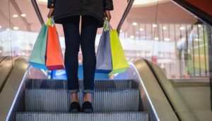 Pessoa subindo escada rolando com sacolas nas mãos em shopping, alusivo às ações da Multiplan