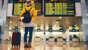 Pessoa de costas com mala de carrinho olhando o celular e o painel de voos, alusivo às viagens que beneficiam ações de Airbnb