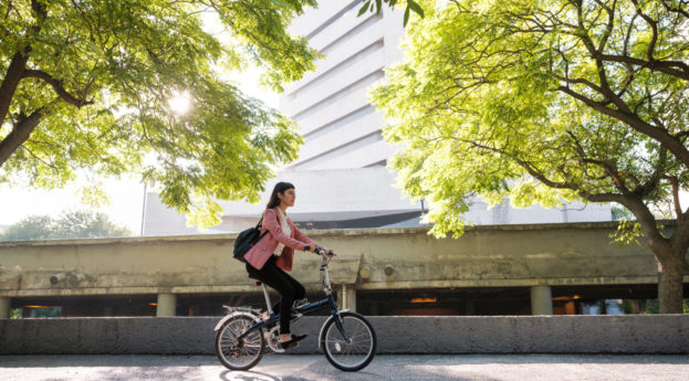 Mulher andando de bicicleta em rua ensolarada com árvores em destaque, alusivo ao conceito ESG