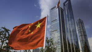 Bandeira da China em primeiro plano com prédios espelhados atrás, alusivo à incorporadora Evergrande e sua venda de fatia em banco