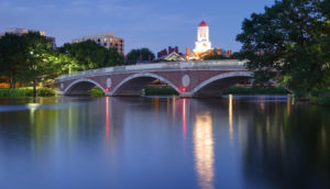 Paisagem da Universidade Harvard, a noite, nos Estados Unidos, com destaque para a torre vermelha e branca iluminada ao fundo e o rio Charles