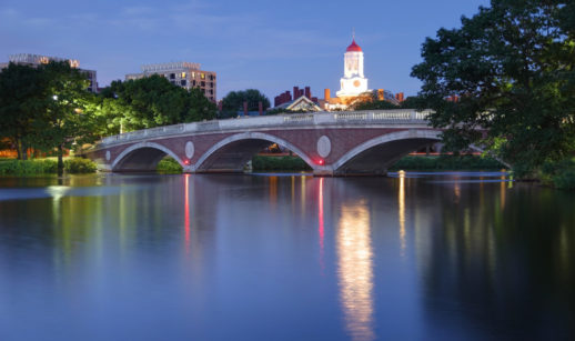 Paisagem da Universidade Harvard, a noite, nos Estados Unidos, com destaque para a torre vermelha e branca iluminada ao fundo e o rio Charles