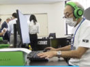 Um dos idosos integrantes da equipe de eSports japonesa Matagi Snipers, mexendo no computador com fone de ouvido grande verde e preto e máscara branca