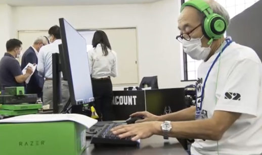 Um dos idosos integrantes da equipe de eSports japonesa Matagi Snipers, mexendo no computador com fone de ouvido grande verde e preto e máscara branca