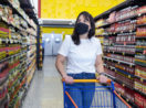 Mulher com carrinho de compras vazio em meio a supermercado olhando os preços de máscara, alusivo à inflação