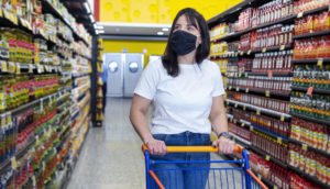 Mulher com carrinho de compras vazio em meio a supermercado olhando os preços de máscara, alusivo à inflação