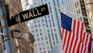 Foto da placa da Wall Street com bandeira dos Estados Unidos ao fundo, alusivo aos índices Nasdaq-100 e S&P-5000