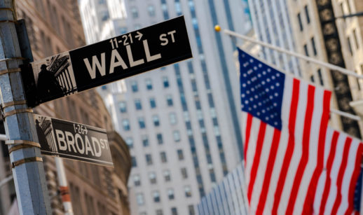 Foto da placa da Wall Street com bandeira dos Estados Unidos ao fundo, alusivo aos índices Nasdaq-100 e S&P-5000