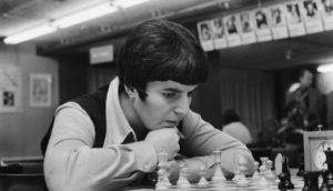 Nona Gaprindashvili, que aparece em O Gambito da Rainha, pensando durante jogo de xadrez, com as mãos no queixo, em preto e branco