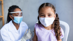 Enfermeira aplicando vacina da Pfizer que está sorrindo com máscara olhando de frente