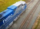 Vista aérea de trem da Rumo, com destaque para o logo da empresa em vagão azul, andando sobre ferrovia