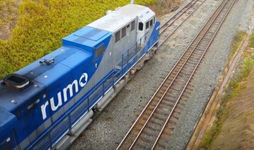 Vista aérea de trem da Rumo, com destaque para o logo da empresa em vagão azul, andando sobre ferrovia