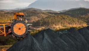 Máquina de mineração com montanha de minério de ferro em primeiro plano e, ao fundo, floresta com montanha, alusivo às ações da Vale (VALE3)
