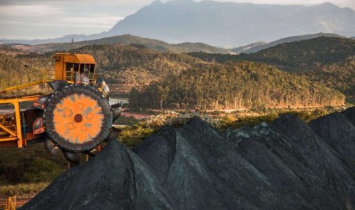 Máquina de mineração com montanha de minério de ferro em primeiro plano e, ao fundo, floresta com montanha, alusivo às ações da Vale (VALE3)