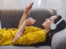 Mulher deitada em sofá com fone de ouvido olhando o celular, alusivo a um vídeo do YouTube
