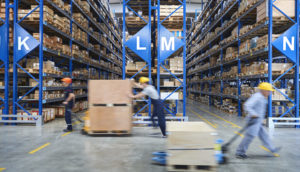 Homens de uniforme azul e capacete amarelo movimentando caixas em meio à armazém, alusivo à atividade econômica do Brasil