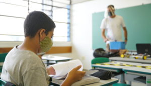 Aluno com máscara e livro nas mãos com professor ao fundo, alusivo às aulas presenciais nas escolas do Estado de São Paulo