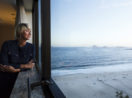 Mulher à direita olhando por janela com vista para a praia no Rio de Janeiro, alusivo à live Conjuntura Safra Invest