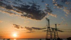 Torres de energia elétrica no lado direito, com linhas de transmissão atravessando a foto e pôr do sol ao fundo