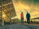 Foto de baixo para cima de funcionários caminhando em campo de energia solar com pôr do sol, alusivo ao fim do uso de combustíveis fósseis por parte dos EUA no setor de energia