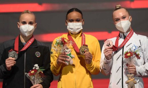Rebeca Andrade no meio de pódio com duas ginastas ao lado, recebendo o ouro