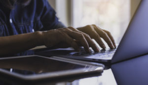 Mãos de pessoa teclando em laptop com tablet ao lado, alusivo ao setor de tecnologia