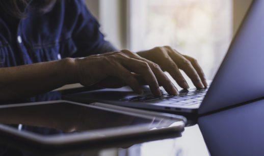 Mãos de pessoa teclando em laptop com tablet ao lado, alusivo ao setor de tecnologia