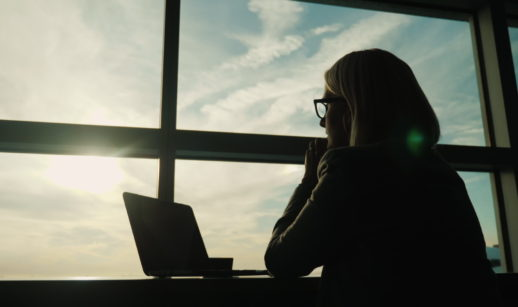Silhueta de mulher sentada com laptop aberto olhando por janela com raio solar entrando, alusivo à espera dos balanços do setor financeiro