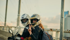 Casal sobre uma motocicleta, de capacete e óculos de sol, com a mulher fazendo sinal de paz e amor com os dedos, alusivo às vendas de motos em setembro