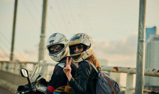 Casal sobre uma motocicleta, de capacete e óculos de sol, com a mulher fazendo sinal de paz e amor com os dedos, alusivo às vendas de motos em setembro