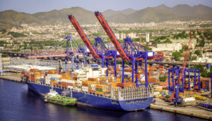 Vista aérea do porto do Rio de Janeiro, com pontes rolantes içando cargas para dentro de navio, alusivo à prévia do PIB da agenda econômica