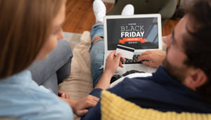 Homem e mulher sentados em sofá com o laptop e cartão de crédito em mãos prontos para comprar na Black Friday